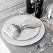 A white plate with a Tabletop Classics by Walco silver napkin ring with a fork and knife on a napkin.