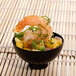 A Fineline black plastic tiny bowl filled with shrimp and pineapple on a bamboo mat on a table.