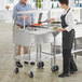 A man and woman standing next to a ServIt stainless steel ice-cooled food table with sneeze guard.