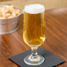 A Libbey stemmed pilsner glass of beer on a table with a napkin and a bucket of peanuts.