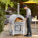 A man wearing an apron cooking a pizza in a Backyard Pro stainless steel wood-fired pizza oven.