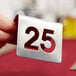 A hand holding a Tablecraft stainless steel cut-out number 25.