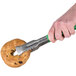 A close-up of a hand holding a bagel with Vollrath VersaGrip tongs with a green handle.