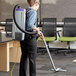 A woman wearing a ProTeam backpack vacuum with a black tube vacuuming a corporate office cafeteria floor.