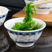 A blue Thunder Group melamine rice bowl filled with green seaweed on a counter with chopsticks.