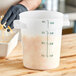 A person in a black glove using a measuring cup to pour white liquid into a large white Carlisle food storage container.