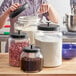 A glass jar with a black lid filled with brown granules on a counter in a professional kitchen.
