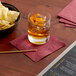 A bowl of potato chips and a drink with an orange peel on a table with a Choice Burgundy cocktail napkin.