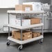 A Metro gray plastic utility cart with three deep ledge shelves holding boxes.