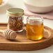 A 1.5 oz. round glass sample jar of honey and a wooden spoon on a table.