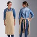 Two women wearing Acopa Ashville natural linen bib aprons over jeans at a counter in a professional kitchen.