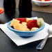 A Carlisle sandshades rimmed fruit bowl filled with fruit on a table with a spoon and a blue bowl of food.