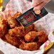 A hand holding a Mike's Hot Honey Extra Hot packet and pouring sauce onto a basket of chicken wings.