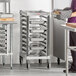 A woman standing in a school kitchen next to a Cambro Camshelving trolley rack with trays on it.