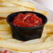 A black oval plastic souffle cup filled with ketchup next to french fries.