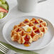 An Avieta individually wrapped pearl sugar Belgian waffle with butter and syrup on a plate next to a bowl of fruit.