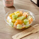 A clear plastic Choice salad bowl filled with sliced fruit with a wooden fork on the table.