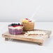 A wooden tray with a bowl of fruit and a bowl of oatmeal with fruit in it, with a white plastic spoon.