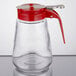 A clear glass Tablecraft syrup dispenser with a red lid.