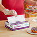 A woman in gloves using white and purple Choice Deli Wrap to put a napkin in a box of doughnuts.