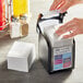 A person's hands using a Tork Xpressnap black tabletop napkin dispenser to take a napkin.