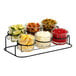 A black metal Cal-Mil 2-tier display with glass jars filled with lemons, limes, cherries, olives, and other food.