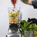 A person pouring grapes into a Breville Commercial blender filled with vegetables and fruits.