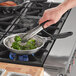 A person cooking broccoli in a Vollrath stainless steel fry pan with a black silicone handle.