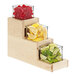 A Cal-Mil maple wood 3-step condiment display with glass containers of lemons and limes on a counter.