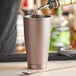 A person using a Barfly antique copper cocktail shaker to pour a drink into a cup.