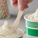 A hand holding a Choice eco-friendly unwrapped wooden taster spoon over a green and white cup of frozen yogurt.