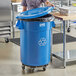 A woman standing in a school kitchen next to a Lavex blue recycling can with a blue lid.