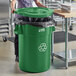 A woman in a school kitchen putting a black garbage bag into a green Lavex recycling can.