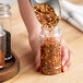 A person pouring red pepper flakes into a round plastic spice jar.