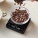 A hand pouring coffee beans into a bowl on a Taylor digital coffee scale.