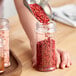 A person pouring red pepper seeds into a round plastic spice jar.