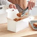 A hand holding a piece of fried chicken in a white barn take-out box.