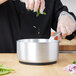 A person pouring greens into a Vollrath Wear-Ever aluminum sauce pan on a counter in a professional kitchen.