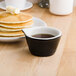 A stack of pancakes with butter and syrup on a white plate with a black and eggshell Tuxton fluted ramekin of liquid.