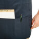 A person's hand holding a pen and a piece of paper in a Chef Revival navy blue bib apron pocket.