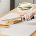 A person in gloves using a Dexter-Russell serrated utility knife to cut bread on a cutting board.
