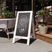 A Flash Furniture Canterbury vintage whitewashed wood A-frame chalkboard sign on a sidewalk.
