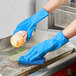 A person wearing blue Showa 707D gloves pouring yellow liquid onto a sponge on a kitchen counter.