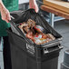A woman putting an EcoStrong Plus black trash bag into a trash can full of food.