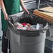 A woman in a green apron holding a black EcoStrong Plus trash bag over a garbage can.