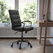A Flash Furniture Whitney black leather office chair with wheels at a desk with a laptop and a book.