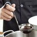 A chef holding a Vollrath stainless steel ladle over a pot of sauce.