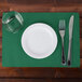 A white plate with a glass and silverware on a Hunter green scalloped paper placemat.
