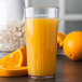 A Carlisle clear plastic tumbler filled with orange juice on a white background next to oranges and a slice of orange.