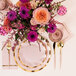 A table setting with Sophistiplate Everyday Blush paper cocktail napkins, gold plates, and flowers.